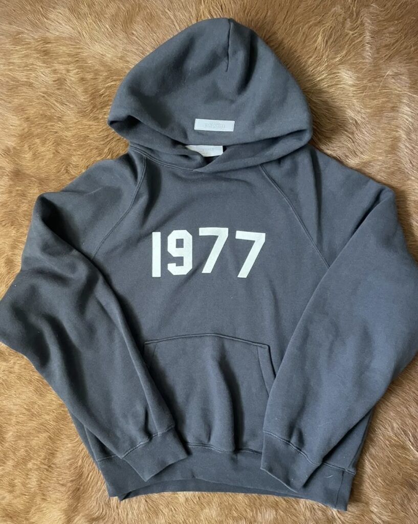 Fear of God Essentials 1977 Hoodie in Iron colorway, featuring bold chest text and minimalist oversized silhouette